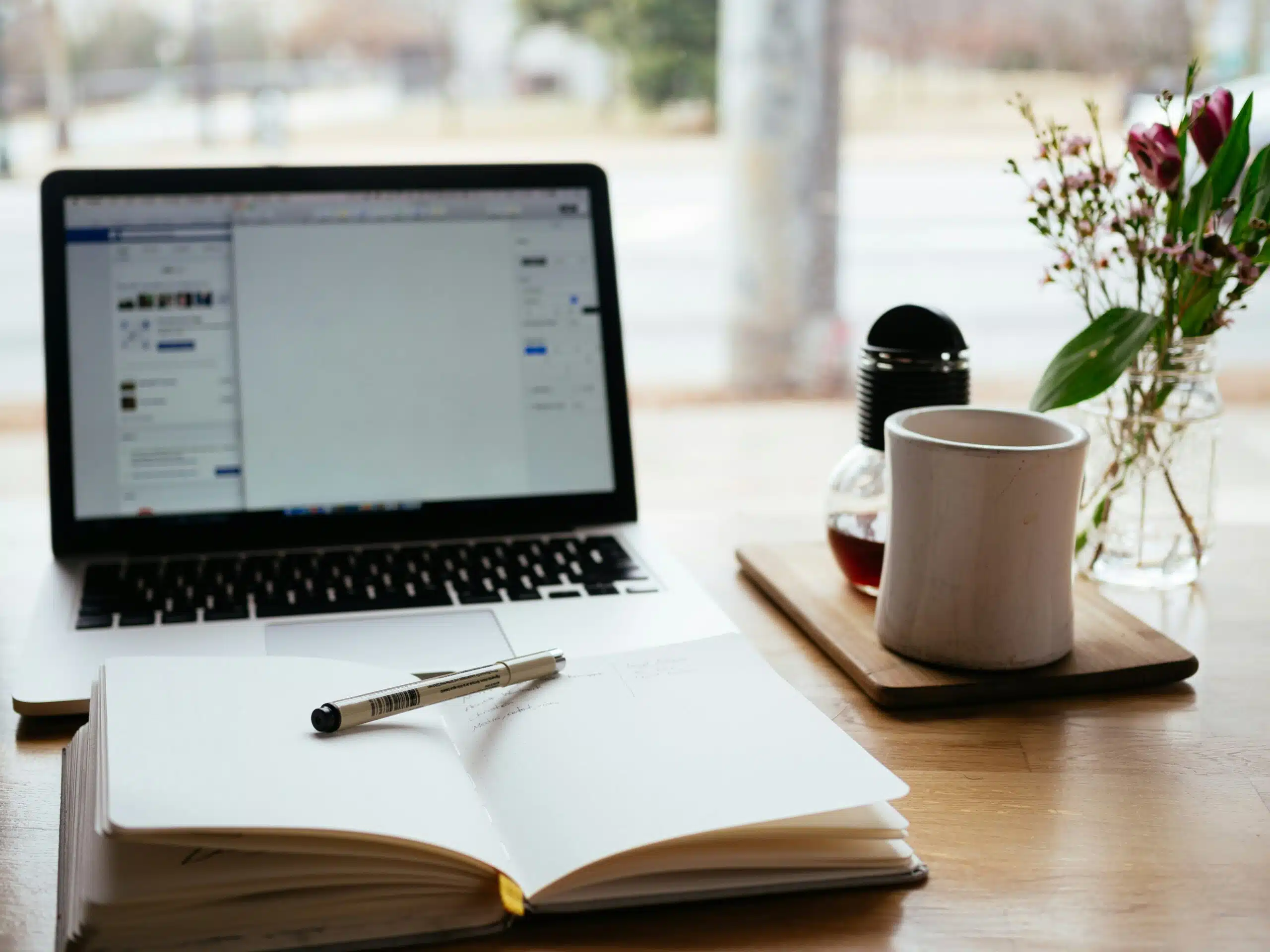 Laptop on a desk with an open notebook, pen, and coffee