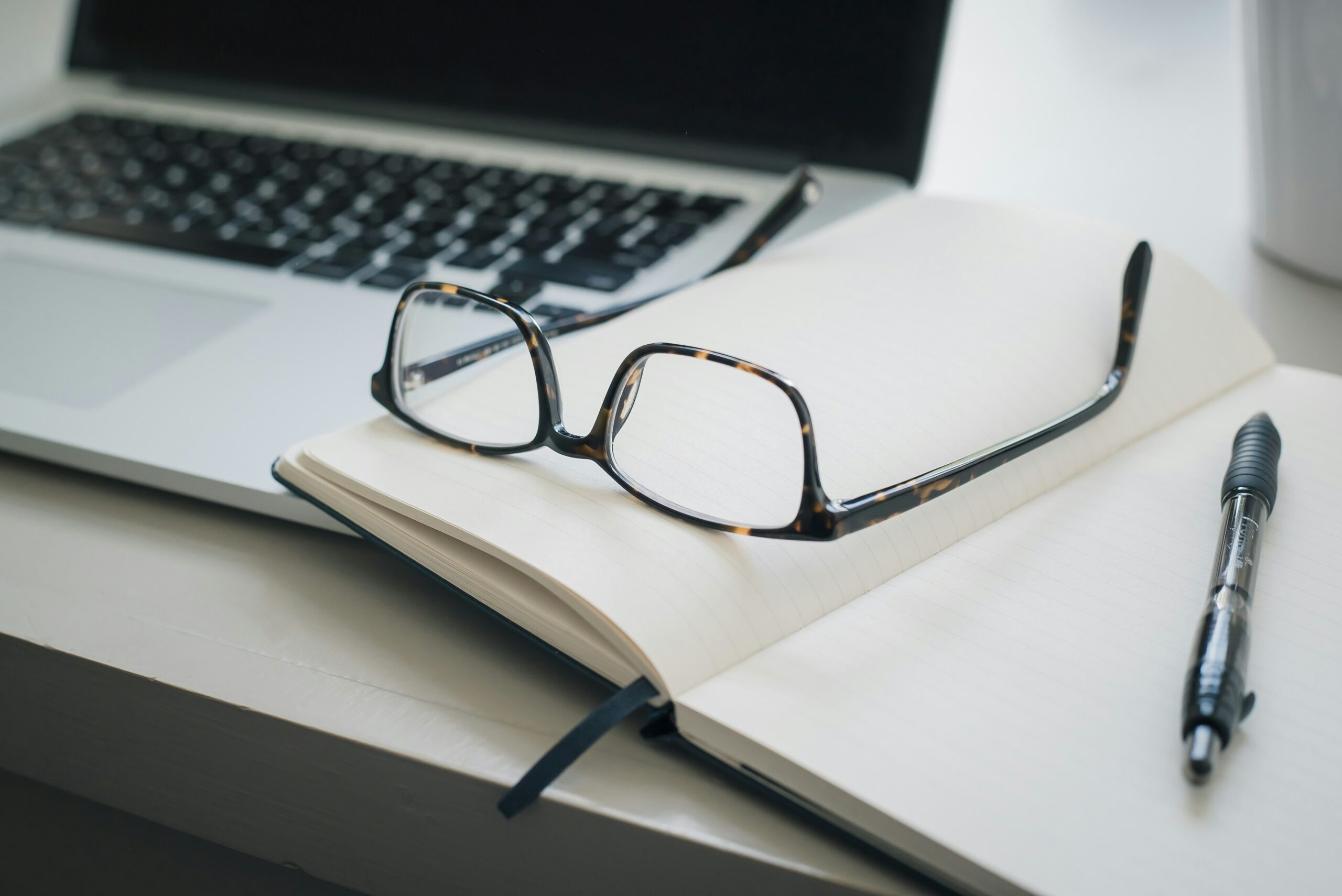 Open notebook with eyeglasses and a pen next to a laptop