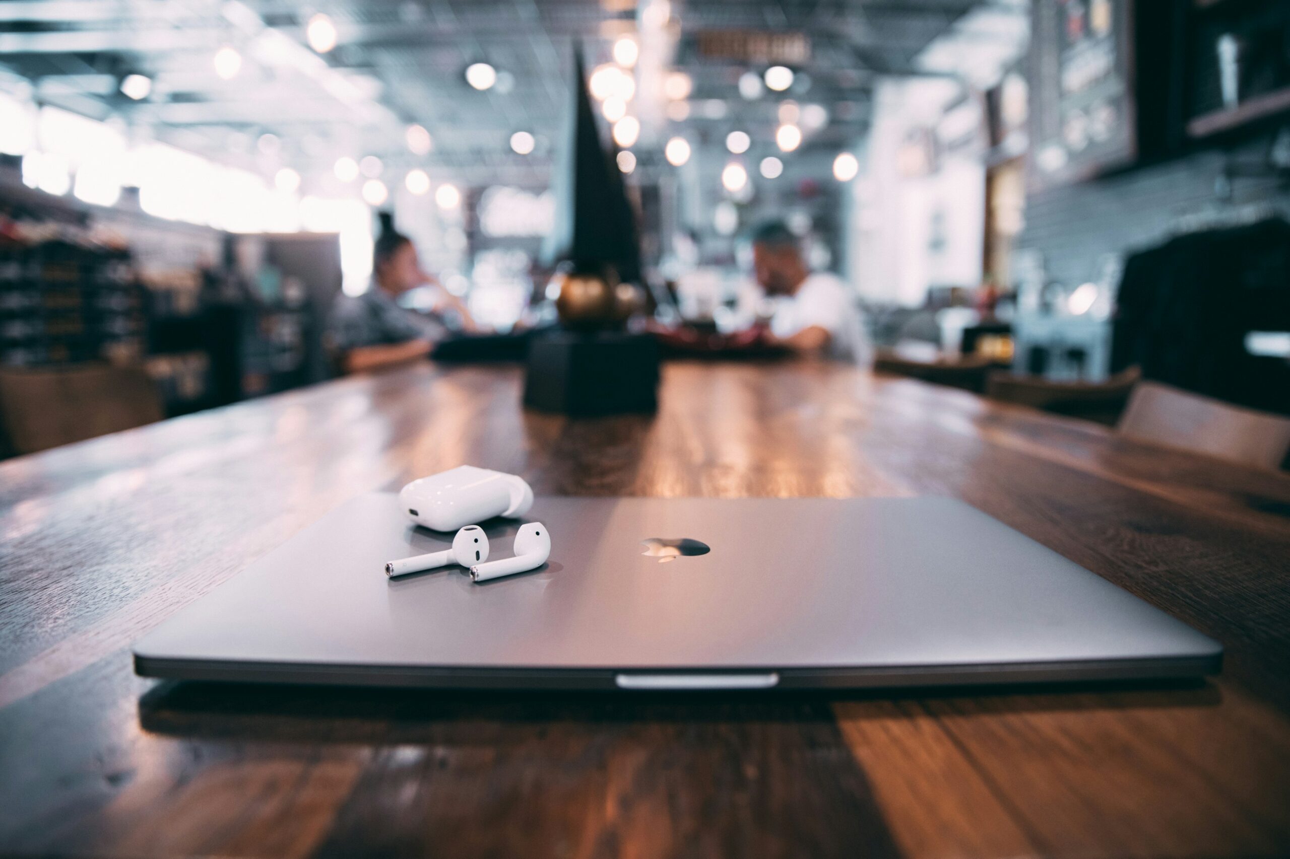 A closed laptop with wireless earbuds on a wooden table in a modern café