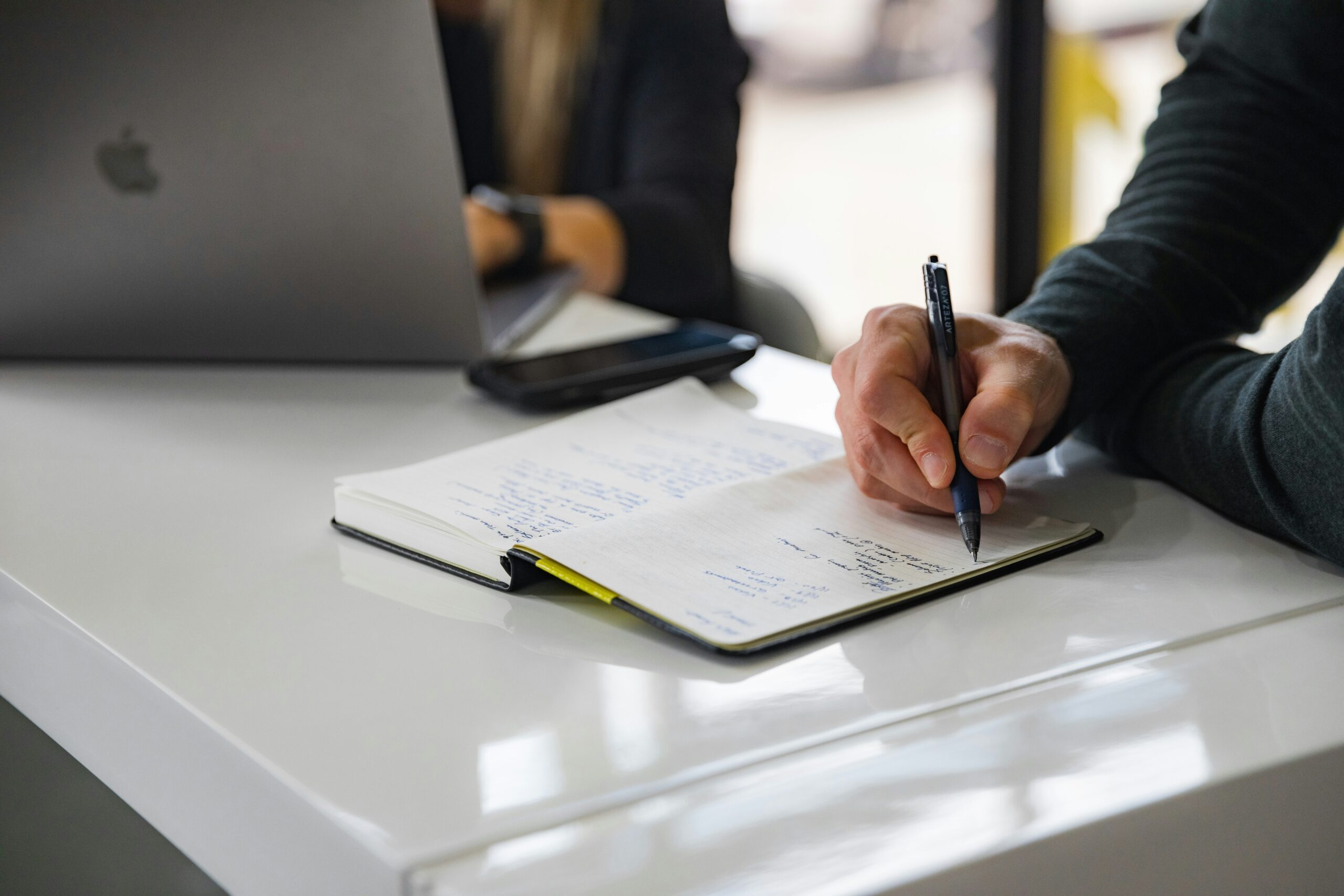 A close-up of a person taking notes in a notebook during a business meeting with a laptop in the background.