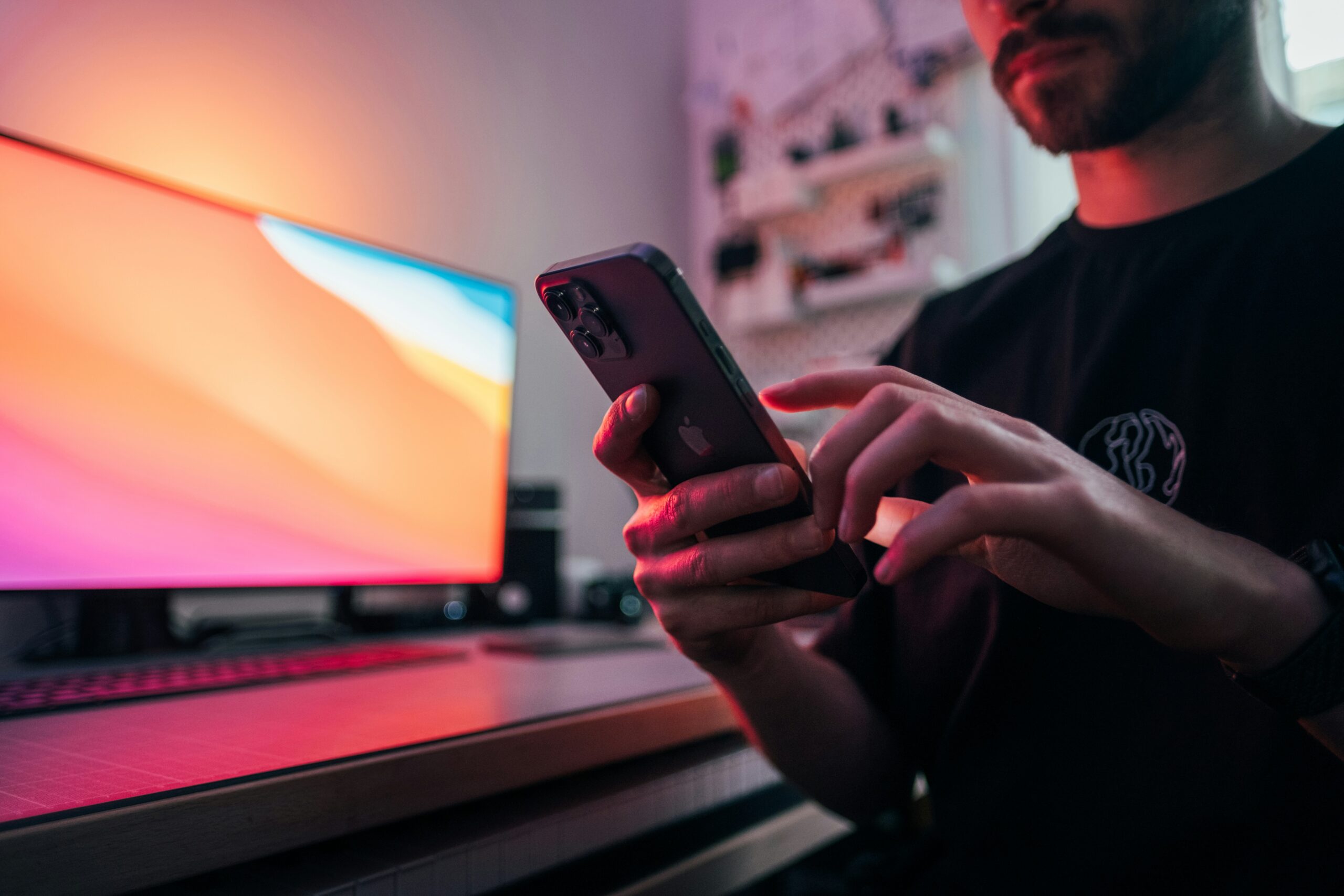 Man using a smartphone in a dimly lit workspace with a glowing desktop monitor in the background.