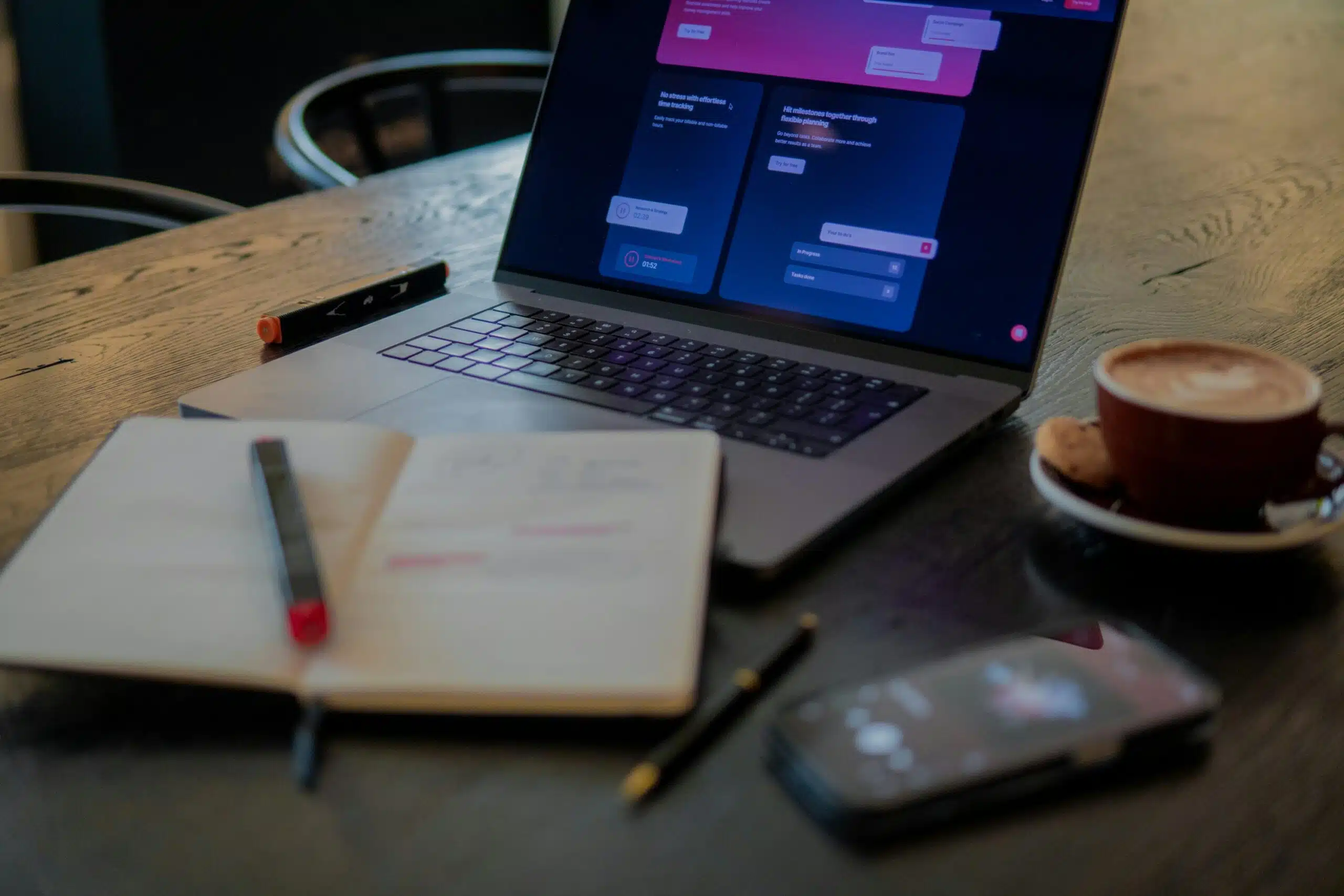 a laptop and a note book set on a desk with pens and a cellphone near by