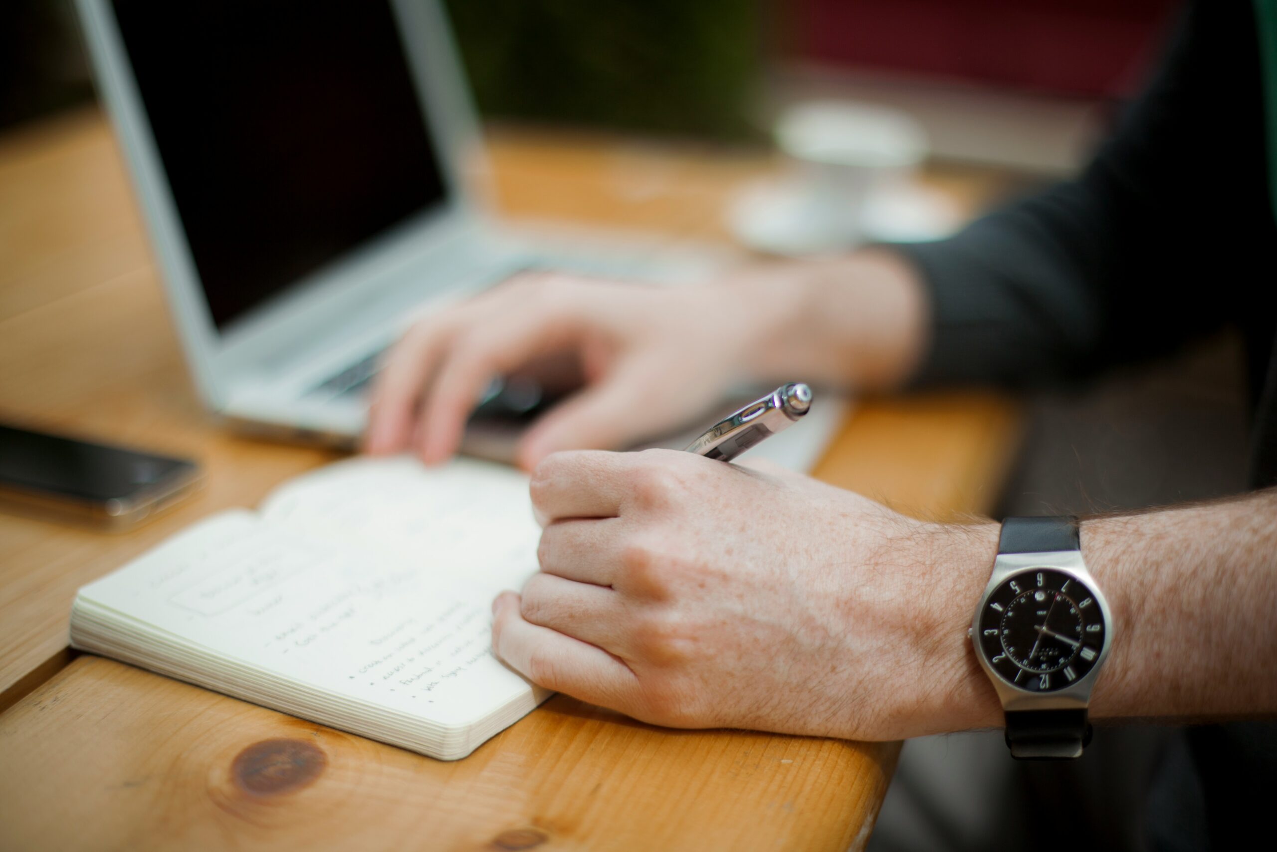 Person taking notes with a pen beside a laptop on a wooden desk.