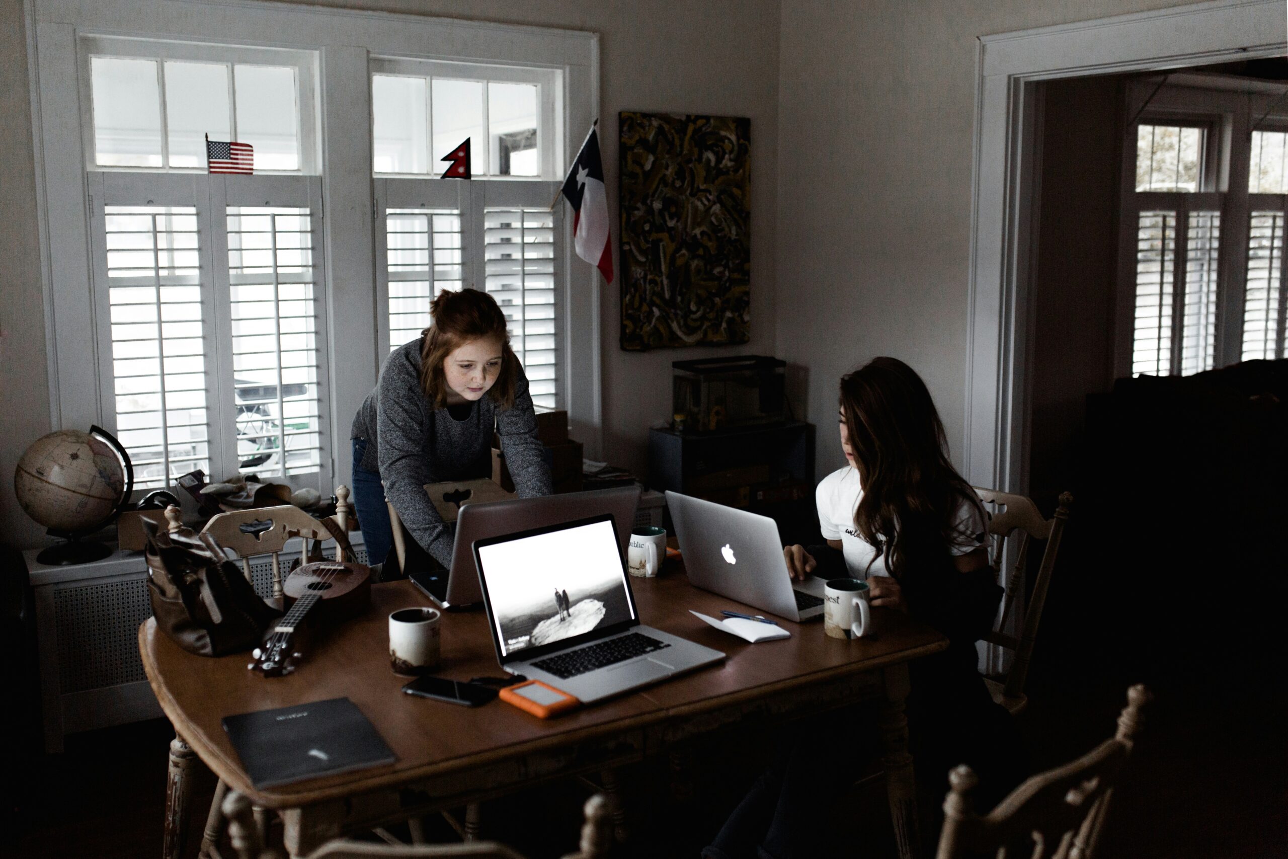 Two women collaborating on laptops in a home office setup with coffee mugs and tech gear. - AI Marketing Tools Two women collaborating on laptops in a home office setup with coffee mugs and tech gear.