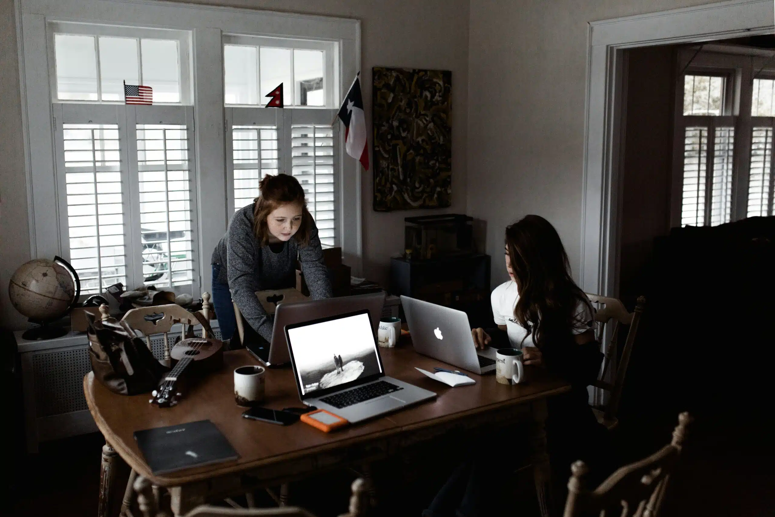 Two women collaborating on laptops in a home office setup with coffee mugs and tech gear.