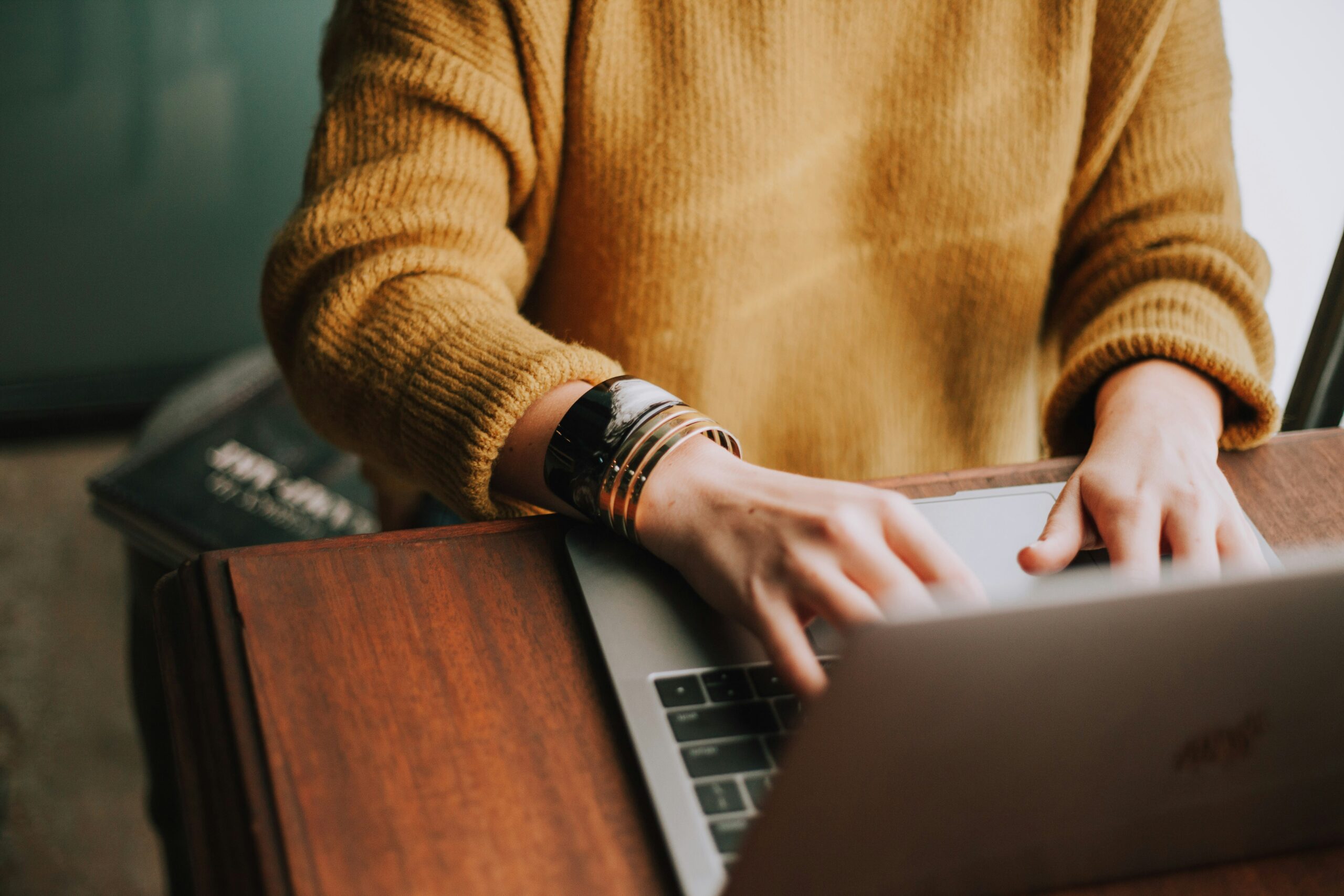 Person in a mustard sweater typing on a laptop at a wooden desk. - AI Marketing Tools Person in a mustard sweater typing on a laptop at a wooden desk.