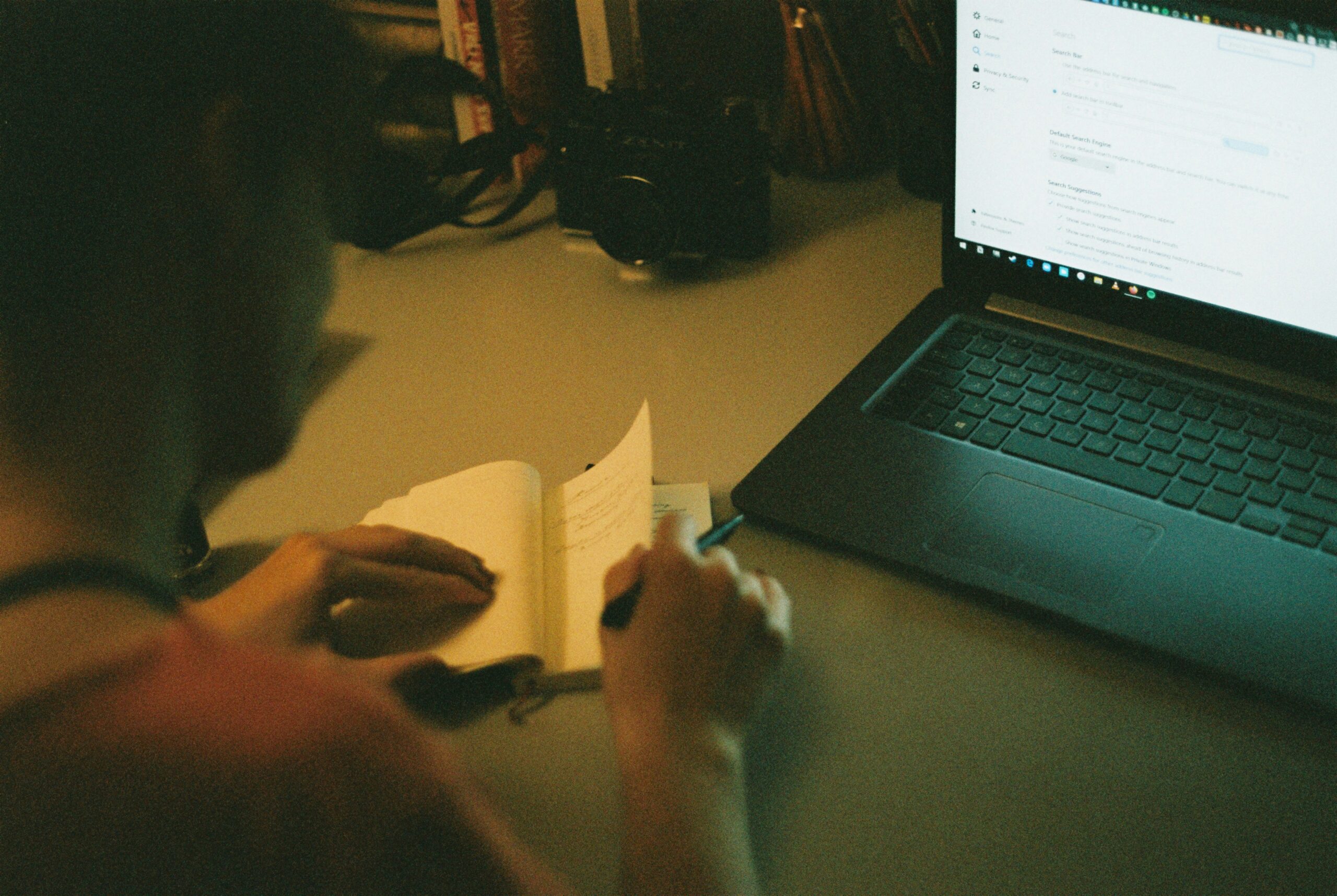 Individual writing in a notebook while referencing a screen with digital tools.