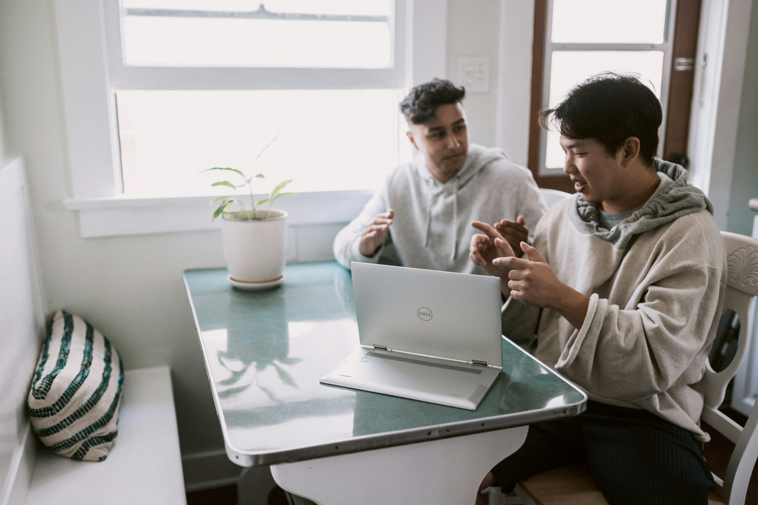 Two men in hoodies having a casual discussion with a laptop on a kitchen table.